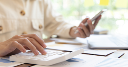 A Male sat at a table, his head is not visible, focus is on his hands. his left hand is holding a notepad and his other hand is ontop of a calculator that is on the table. He could be working out if he can get a mortgage with bad credit history