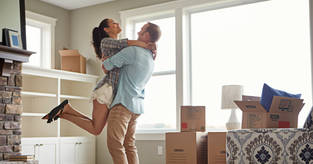 Man and woman in their living room surrounded by packing boxes, they are unpacking, the woman is in the mans arms being lifted up, they are both happy. It could be suggested that they were able to purchase this new home through a shared ownership mortgage.