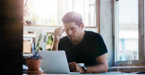 A man sat at a table, using his laptop. he is looking a little stressed and confused, this could be because he doesn't understand remortgaging and is searching about it on his laptop.